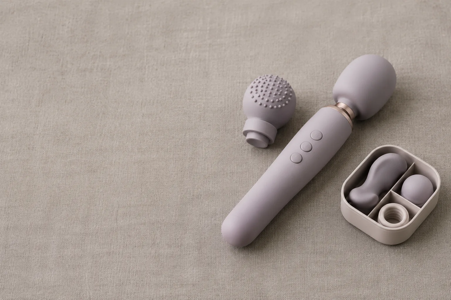 A soft-lit still life on warm gray linen: a matte wand massager, its spare head and a small open parts tray grouped in the right half of the frame, nothing lett